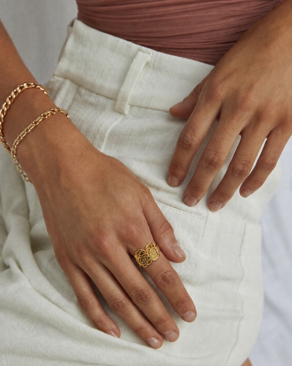 Mão feminina com anel dourado de rosas, pulseiras douradas, calças brancas e top castanho.