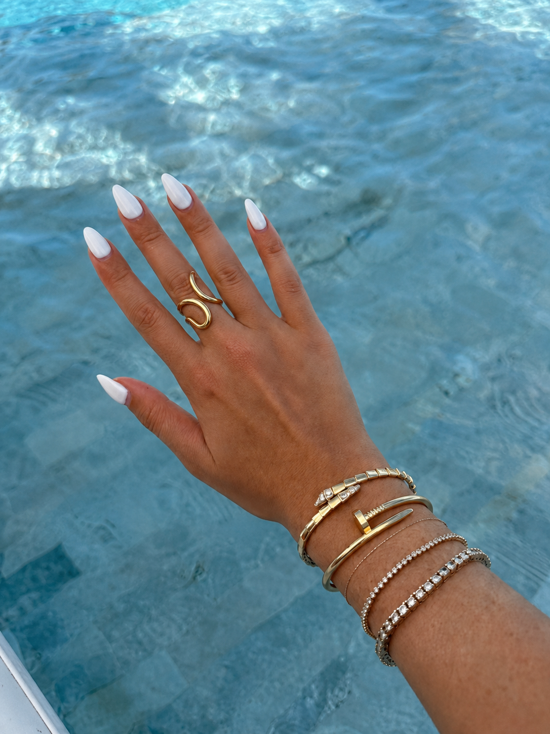 Mão com unhas brancas e várias joias douradas junto a uma piscina
