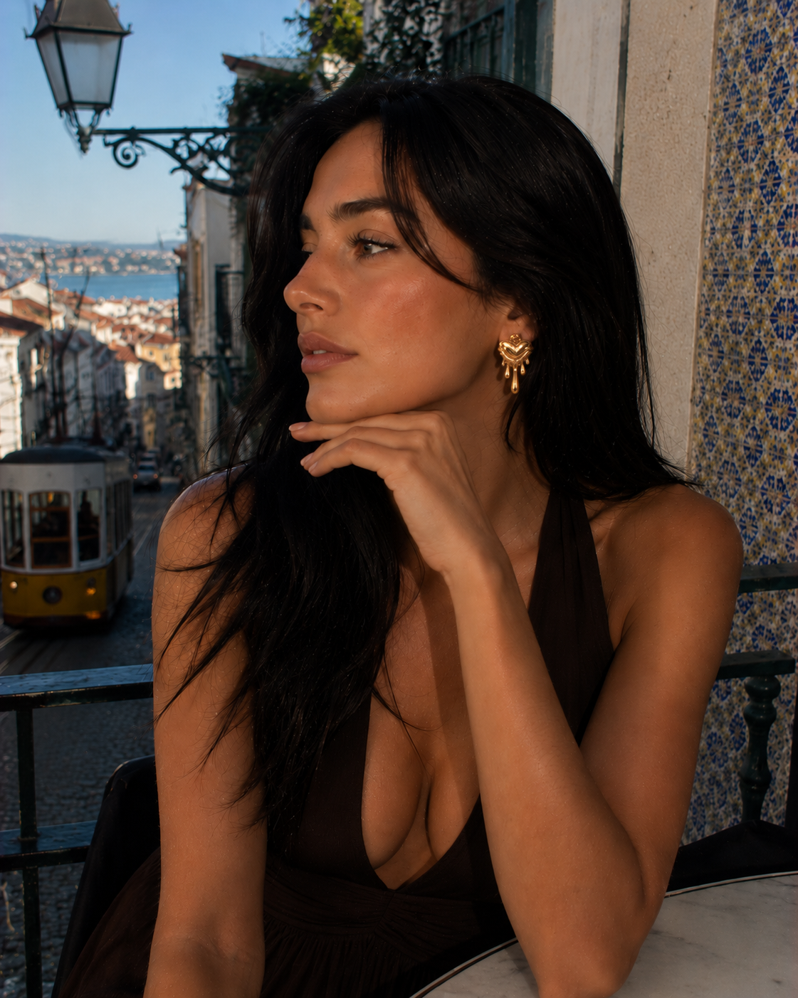 Mulher com brincos dourados em forma de coração, cabelo preto, vestido castanho e azulejos portugueses no fundo