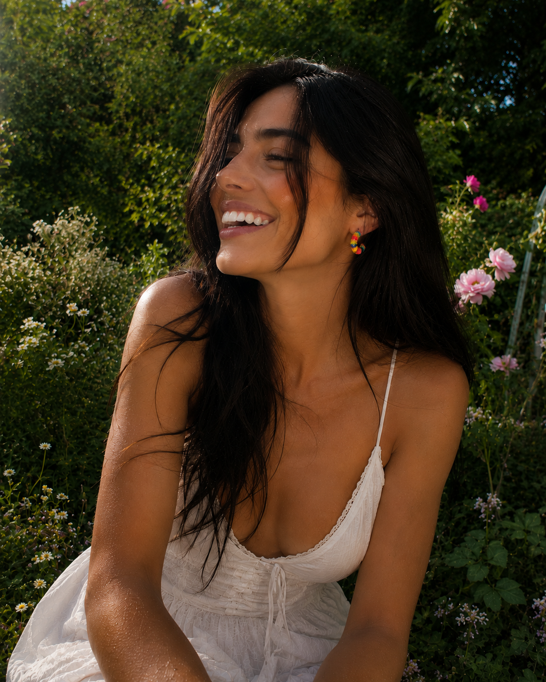 Mulher sorridente de vestido branco e brincos coloridos, sentada num jardim com flores e vegetação verde