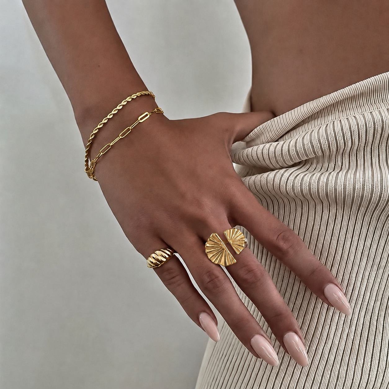 Mão feminina com anéis e pulseiras douradas e saia bege canelada