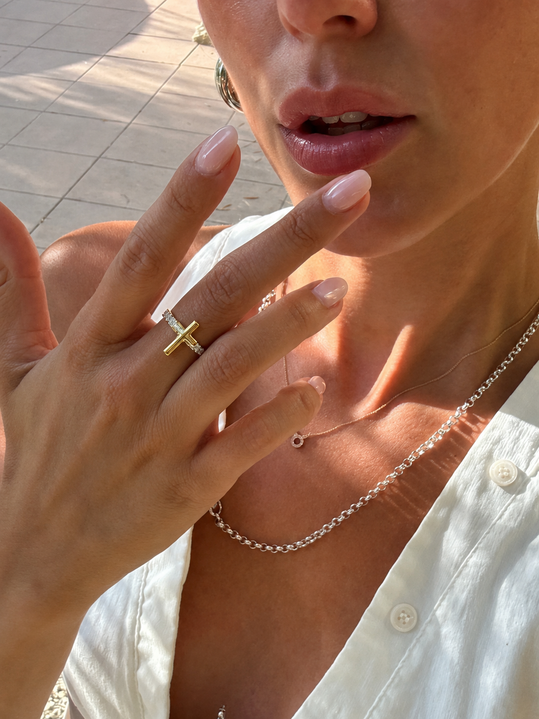 Mão feminina com anel de cruz dourada e colares prateados, unhas nude, blusa branca.