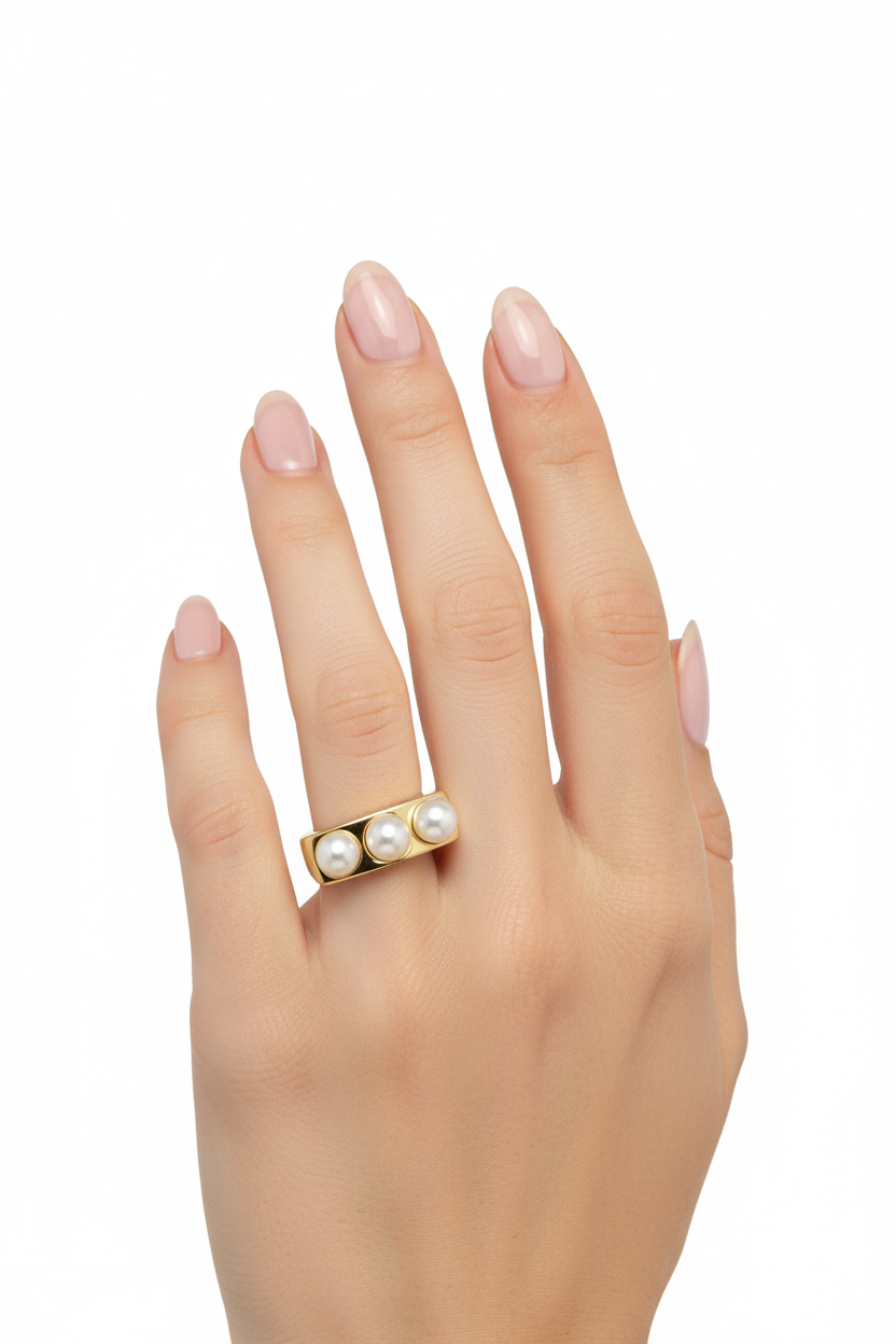 Anel dourado com três pérolas num dedo de mão com unhas naturais e fundo branco