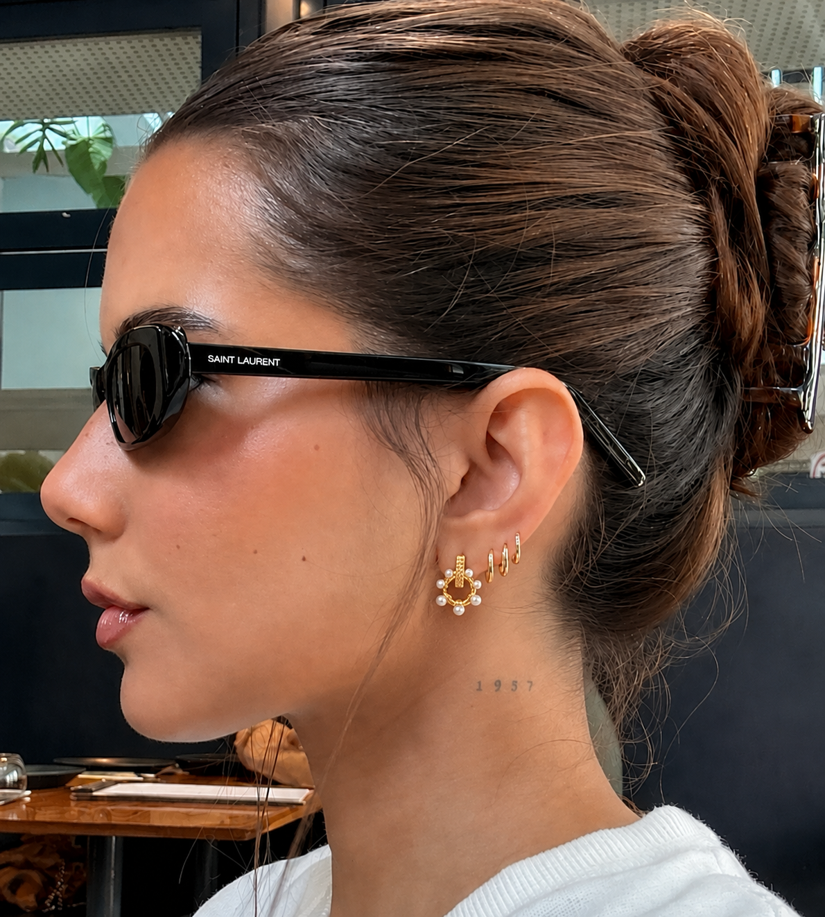 Óculos de sol pretos Saint Laurent em modelo feminino com brincos dourados e tatuagem no pescoço