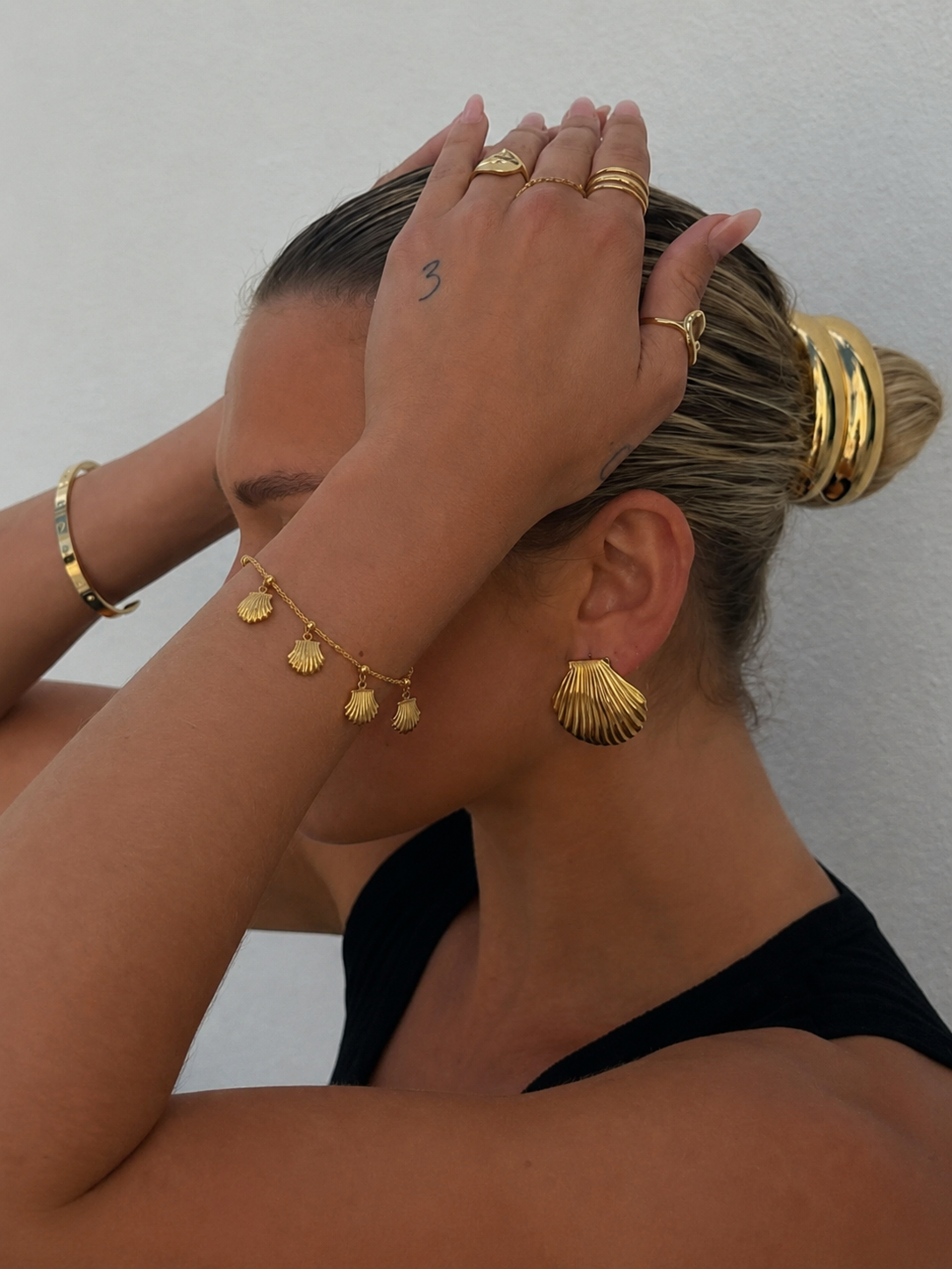 Mulher a usar vários acessórios de joalharia dourada incluindo anéis, pulseira, bracelete rígido, brincos de concha e acessórios de cabelo dourados