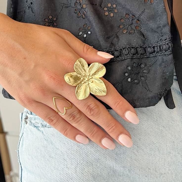 Mão feminina com dois anéis dourados, uma flor grande e um coração, com blusa preta e jeans claros