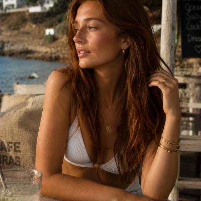 Mulher com biquíni branco e joias douradas sentada numa mesa junto ao mar