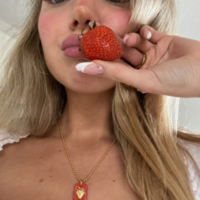 Mulher loira segura morango junto aos lábios e usa colar com pendente vermelho e dourado em forma de morango