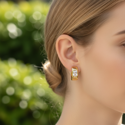 Brinco de ouro amarelo largo com pedra transparente quadrada no centro, usado por mulher de cabelo loiro apanhado.