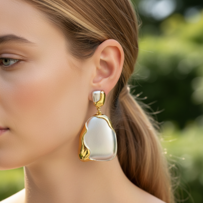 Brinco pendente dourado e prateado com textura metálica num fundo de vegetação