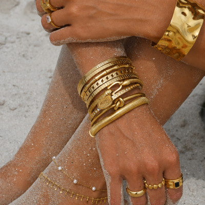 Pulseiras, anéis e tornozeleiras douradas com pérolas em pele bronzeada com areia