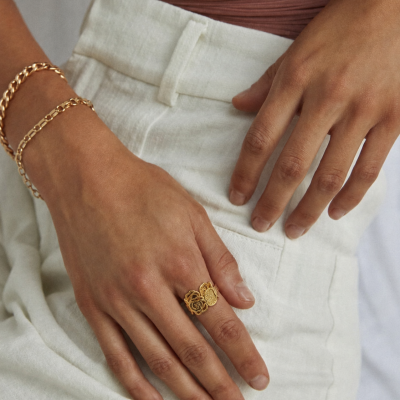 Mão feminina com anel dourado de rosas, pulseiras douradas, calças brancas e top castanho.