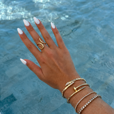 Mão com unhas brancas e várias joias douradas junto a uma piscina