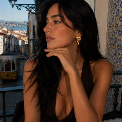 Mulher com brincos dourados em forma de coração, cabelo preto, vestido castanho e azulejos portugueses no fundo