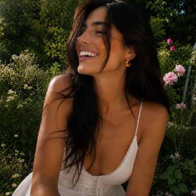 Mulher sorridente de vestido branco e brincos coloridos, sentada num jardim com flores e vegetação verde