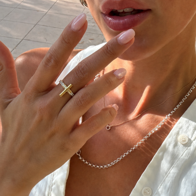 Mão feminina com anel de cruz dourada e colares prateados, unhas nude, blusa branca.