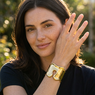 Pulseira dourada larga no pulso de mulher com cabelo castanho e t-shirt preta ao ar livre