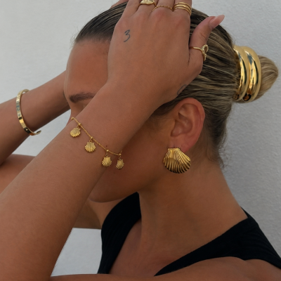 Mulher a usar vários acessórios de joalharia dourada incluindo anéis, pulseira, bracelete rígido, brincos de concha e acessórios de cabelo dourados