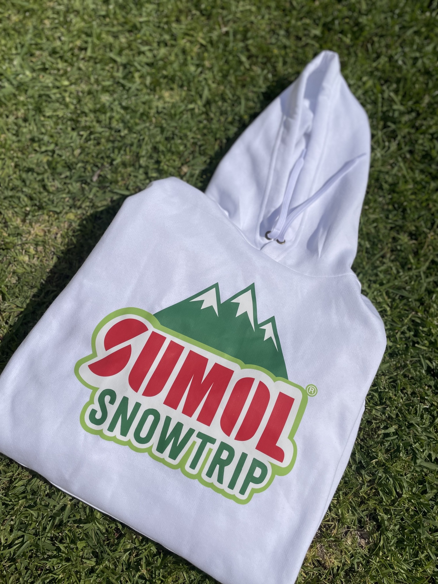 Camisola branca com capuz e estampado colorido com texto SUMOL SNOWTRIP sobre relva verde