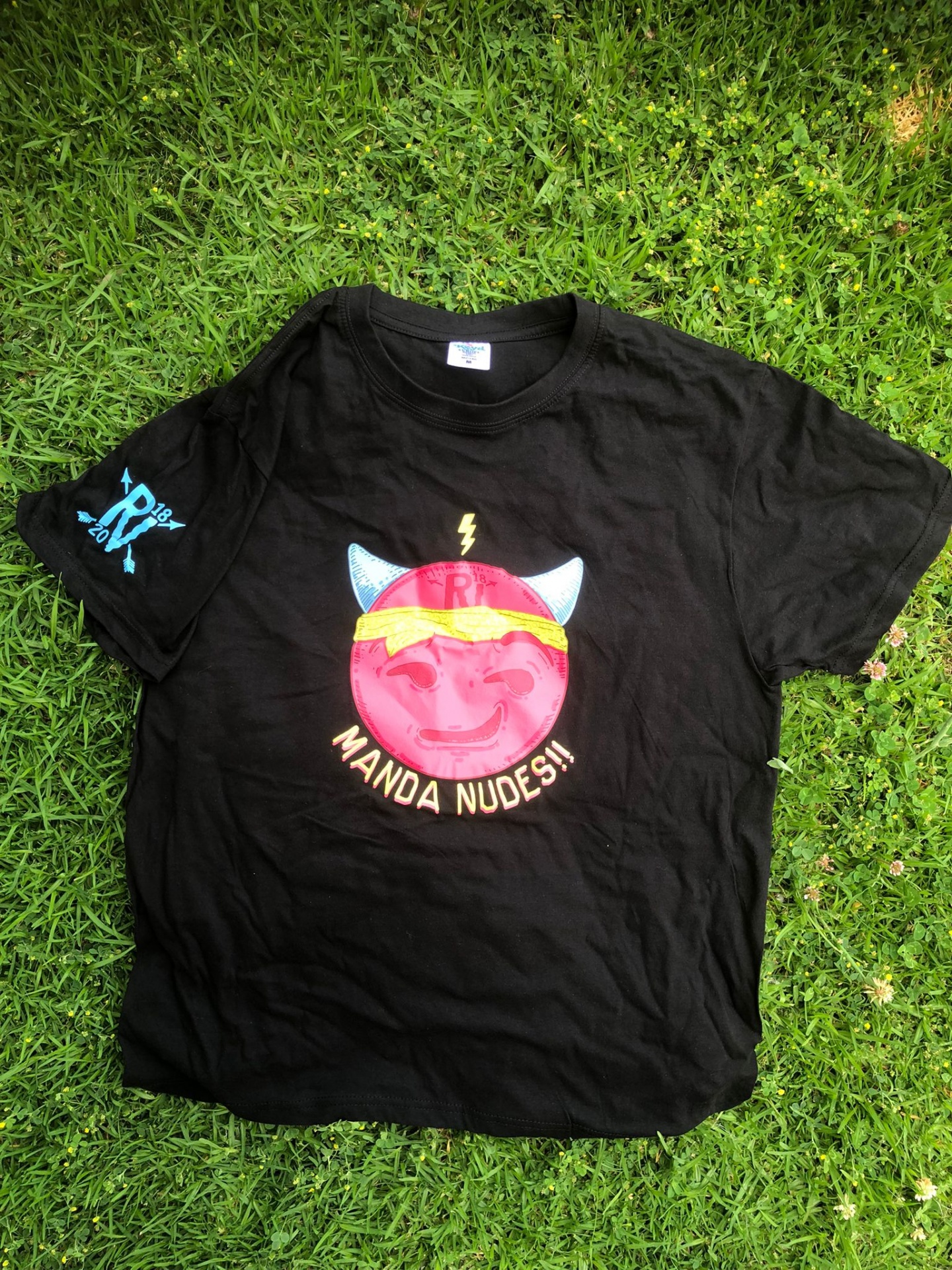 Camisola preta com ilustração colorida de rosto sorridente rosa e texto Manda Nudes no centro, sobre relva verde.