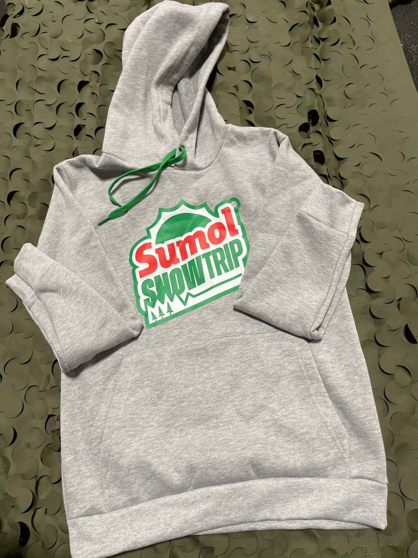 Camisola cinzenta com capuz e cordão verde, estampa 'SUMOL SNOWTRIP' com árvores sobre tecido verde recortado.