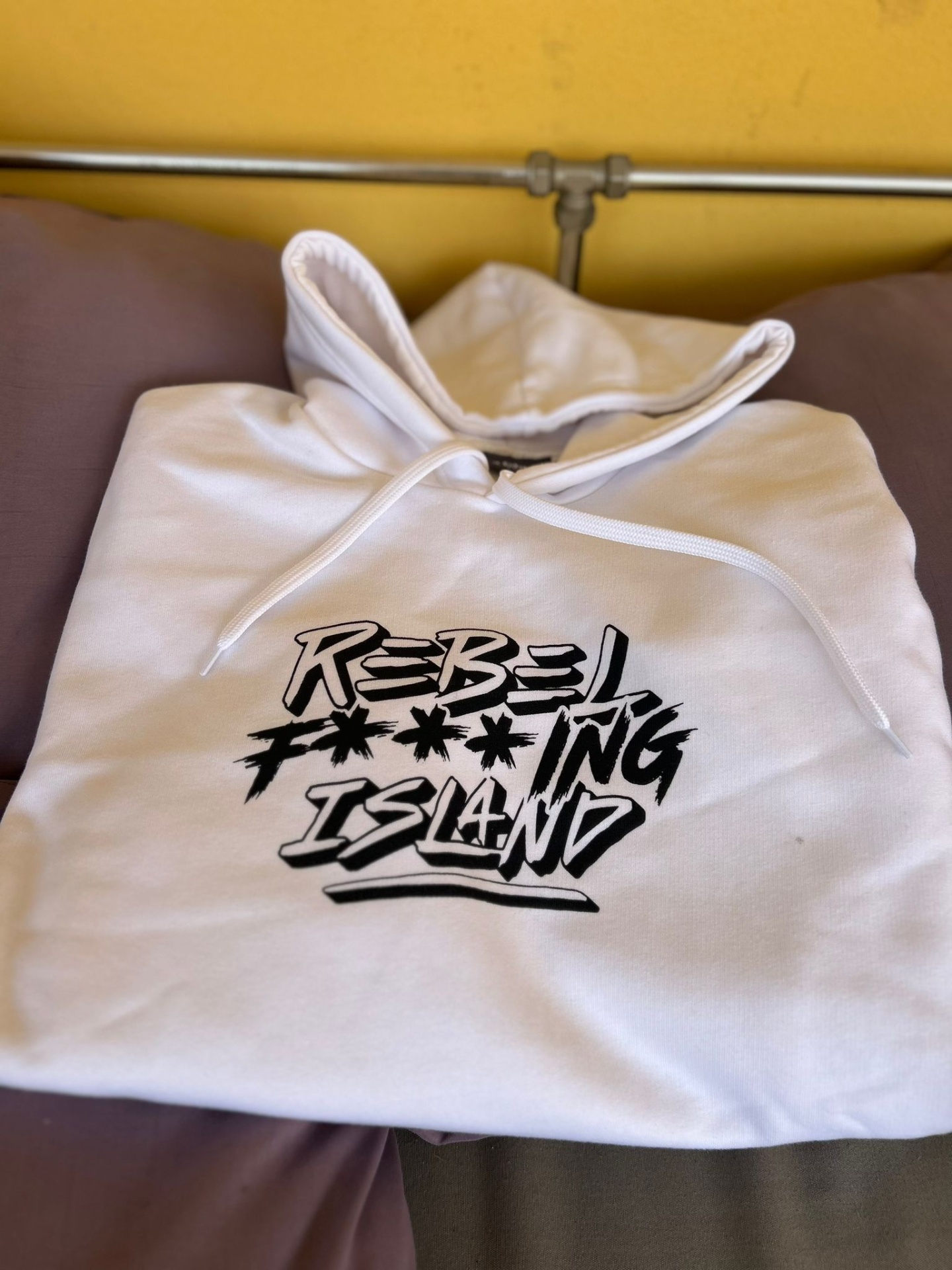 Hoodie branco com capuz e estampa preta REBEL F***ING ISLAND sobre almofadas castanhas e roxas