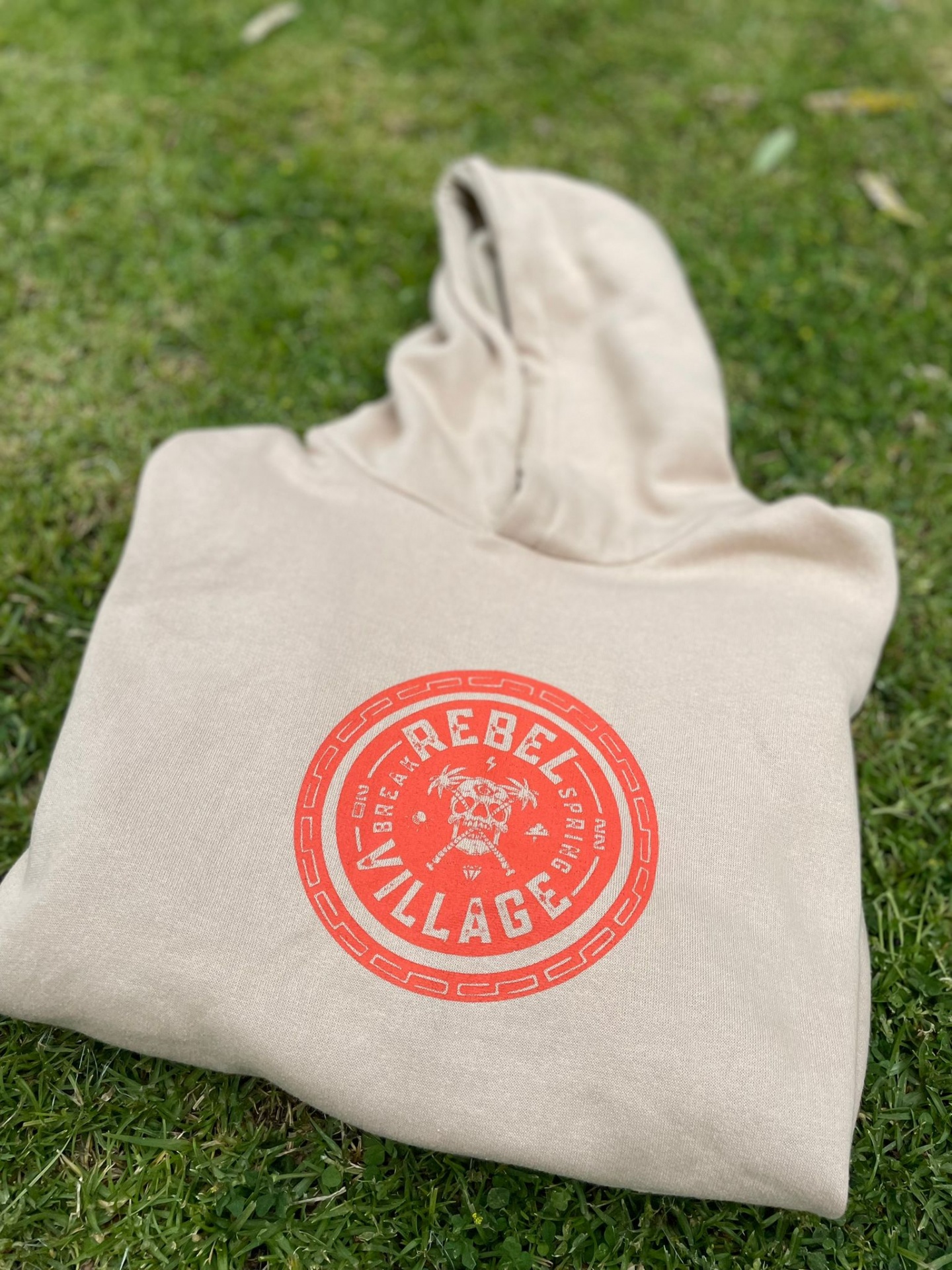 Sweatshirt bege com capuz e estampado vermelho com texto REBEL VILLAGE e tigre, sobre relva verde.