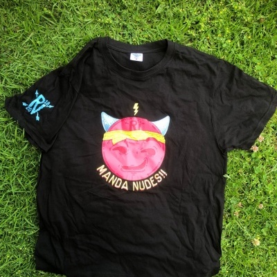 Camisola preta com ilustração colorida de rosto sorridente rosa e texto Manda Nudes no centro, sobre relva verde.