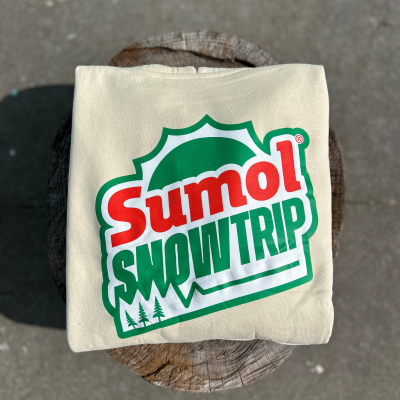 Camisola creme dobrada com estampa Sumol SNOW TRIP em verde e vermelho sobre tronco de madeira