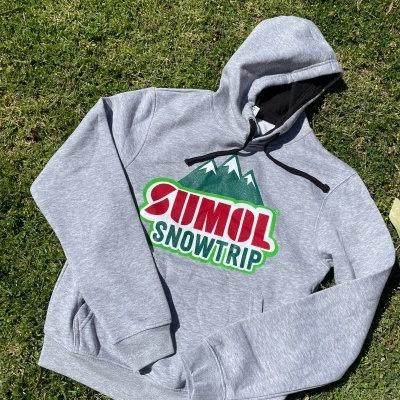 Camisola cinza com capuz e estampa colorida de montanhas e texto SUMOL SNOWTRIP, sobre relva