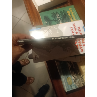 Livro DE NÉ Ó VÍTIMAS DA DÍVIDA segurado, outros livros empilhados sobre móvel de madeira