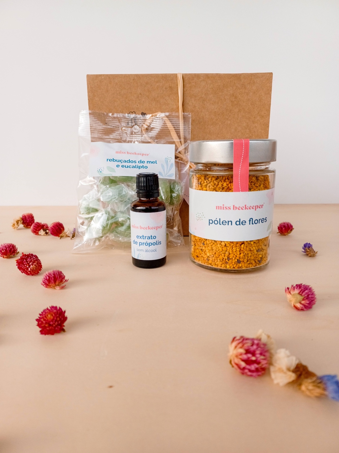 Conjunto de produtos de apicultura com pólen de flores, extrato de própolis e rebuçados de mel e eucalipto, com flores secas ao redor.