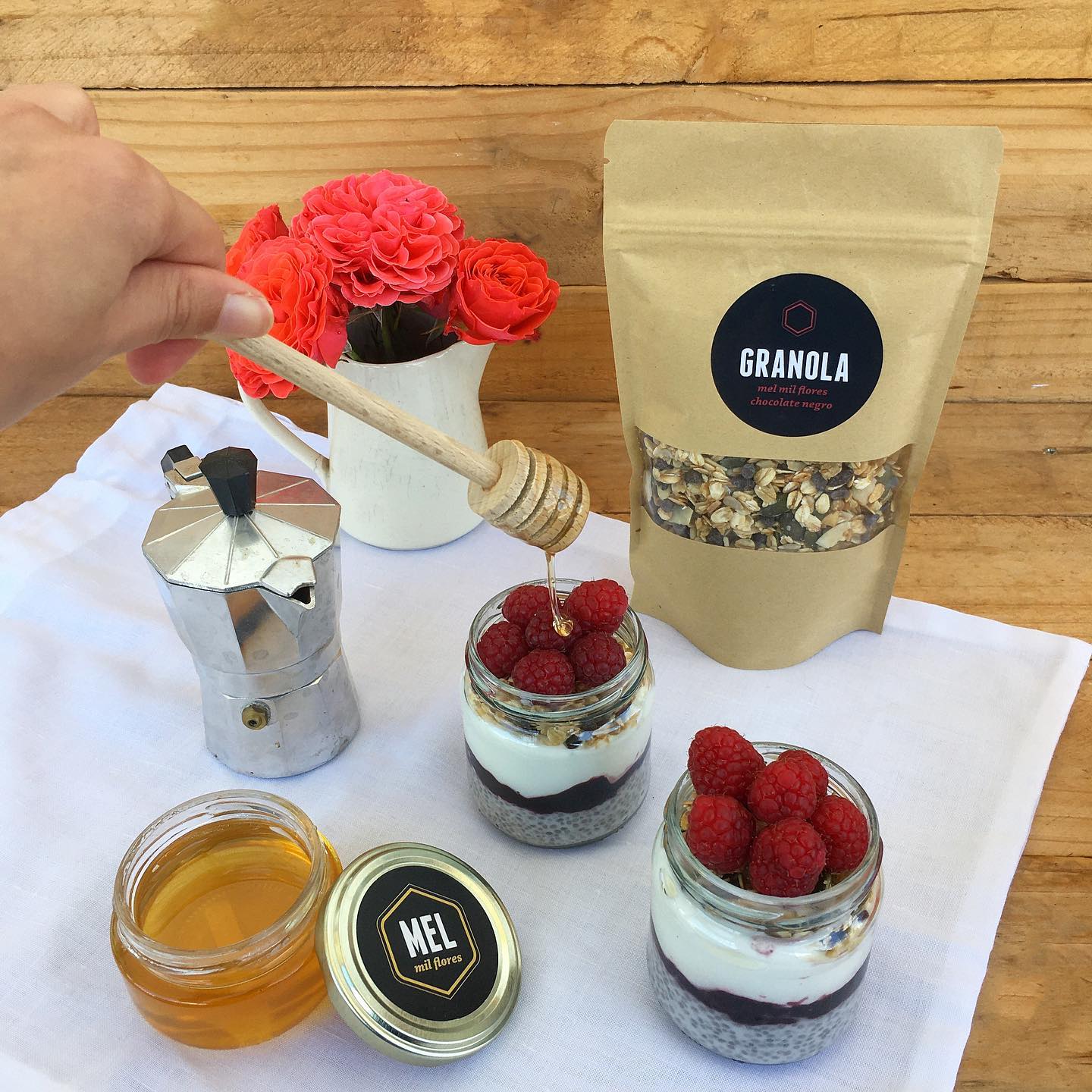 Dois copos de vidro com iogurte, granola e framboesas em cima de pano branco, saco de granola castanho, frasco de mel aberto, cafeteira metálica e vaso branco com flores ao fundo