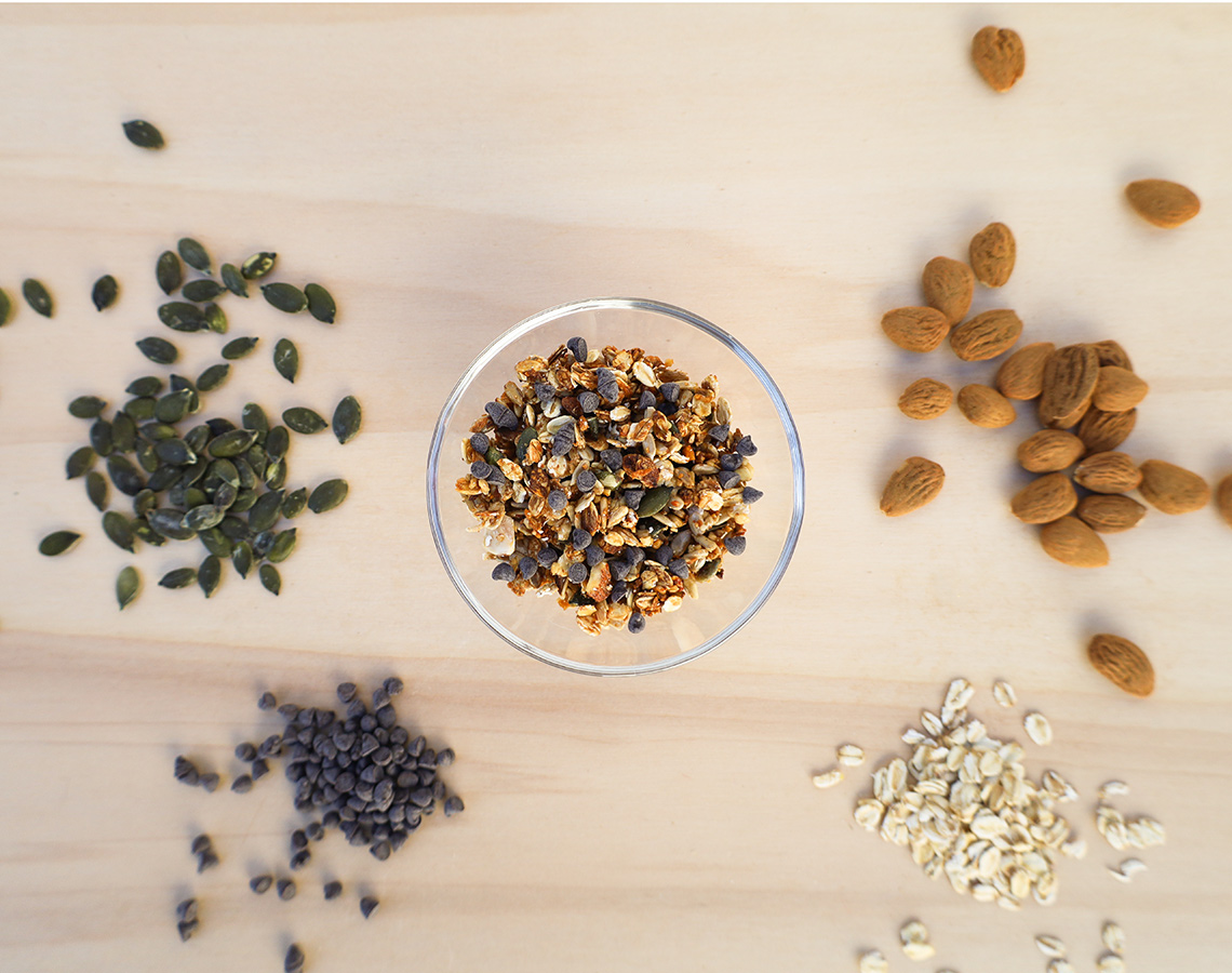 Tigela de vidro com granola cercada por amêndoas, pepitas de chocolate e sementes de abóbora sobre mesa de madeira