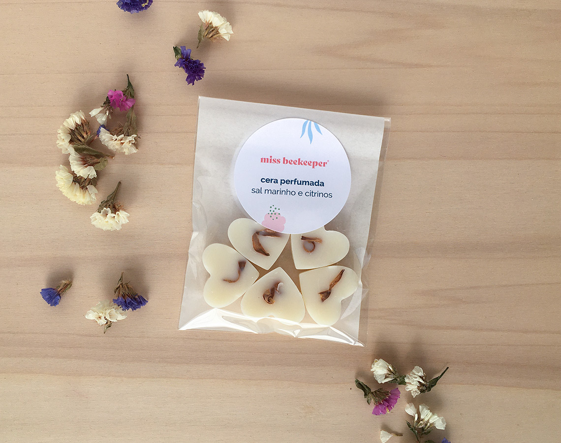 Pacote de corações de cera perfumada com flores secas e etiqueta sobre mesa clara, rodeado de flores secas coloridas.