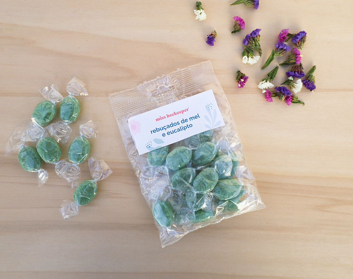Pacote de rebuçados de mel e eucalipto com rebuçados verdes embalados individualmente em madeira clara, flores ao lado.