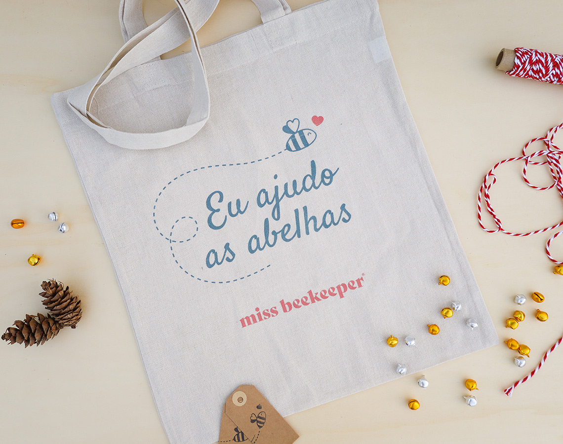 Saco de pano branco com texto e ilustração de abelha, em mesa clara com enfeites e fio