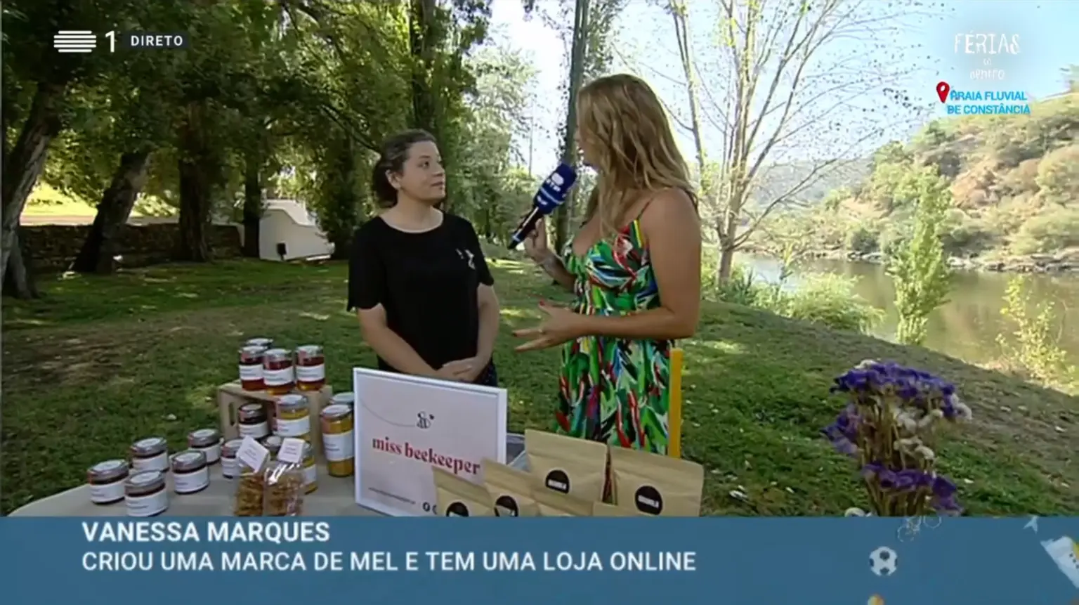 Produtos de mel em frascos num expositor ao ar livre com flores e duas mulheres conversando