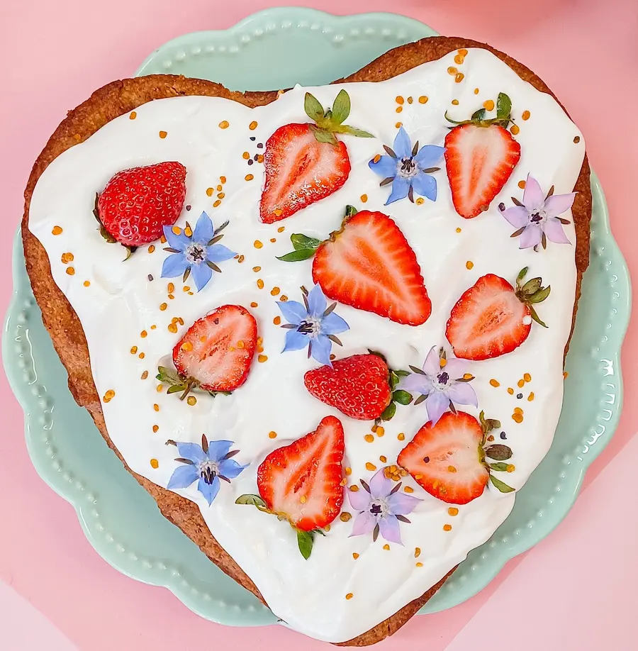 Bolo em forma de coração com cobertura branca e decoração de morangos e flores sobre prato verde claro
