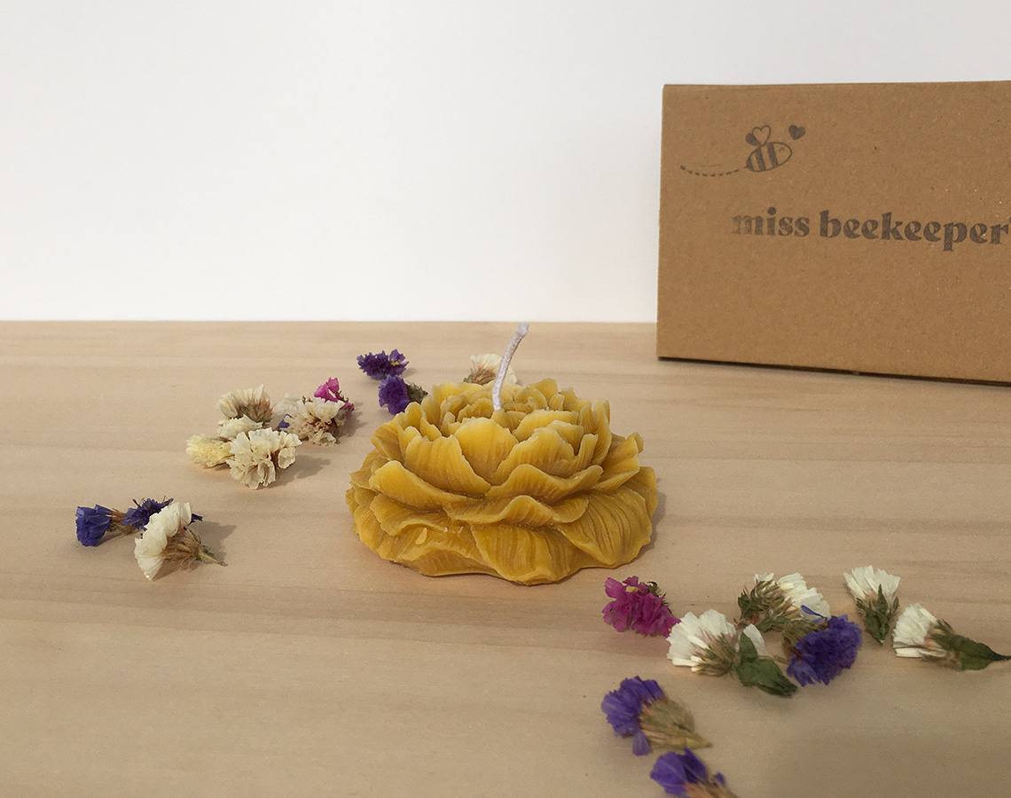 Vela amarela em forma de flor com flores secas ao redor e caixa de papelão ao fundo