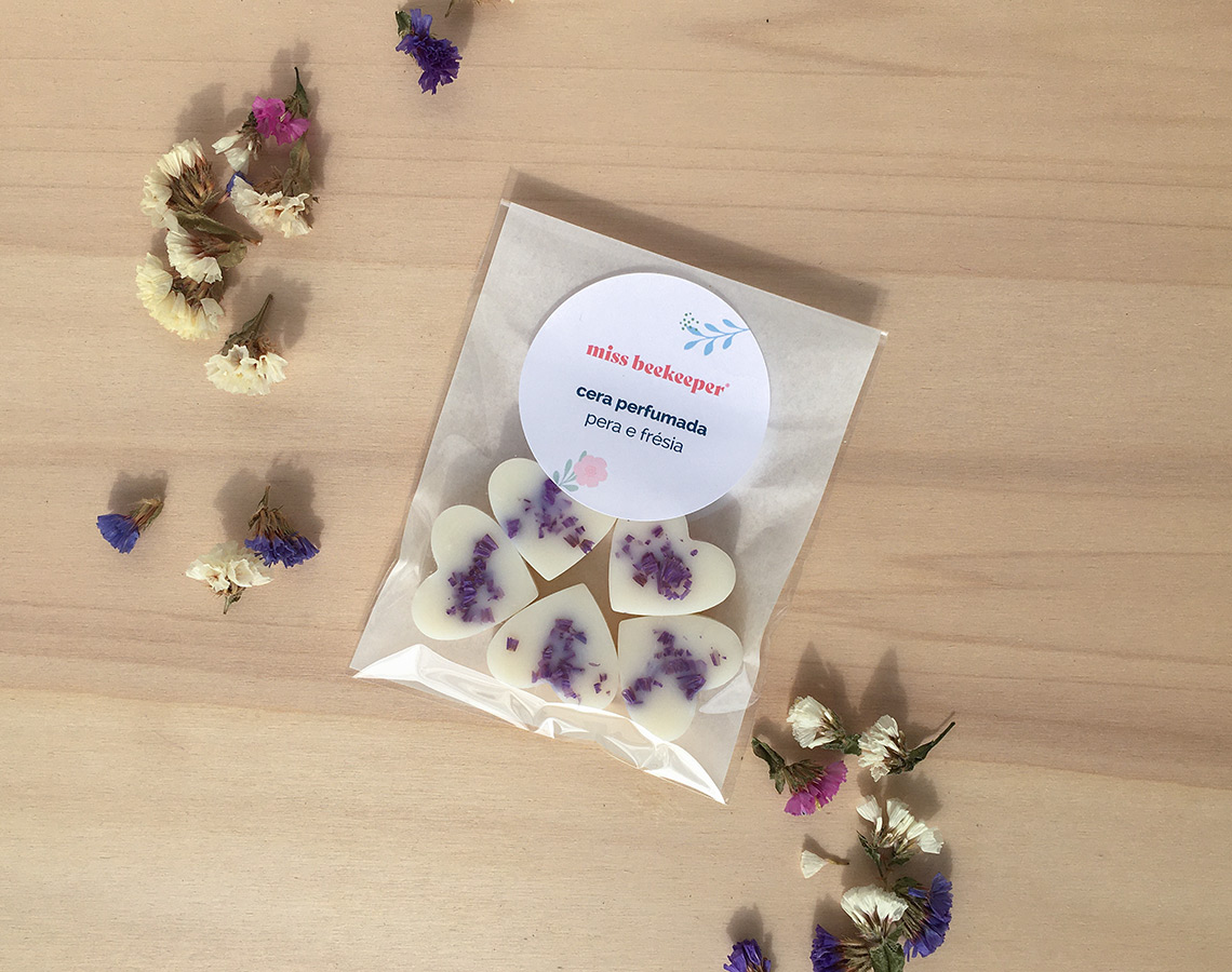 Cinco velas em forma de coração com flores roxas em embalagem transparente sobre mesa de madeira com flores secas ao redor