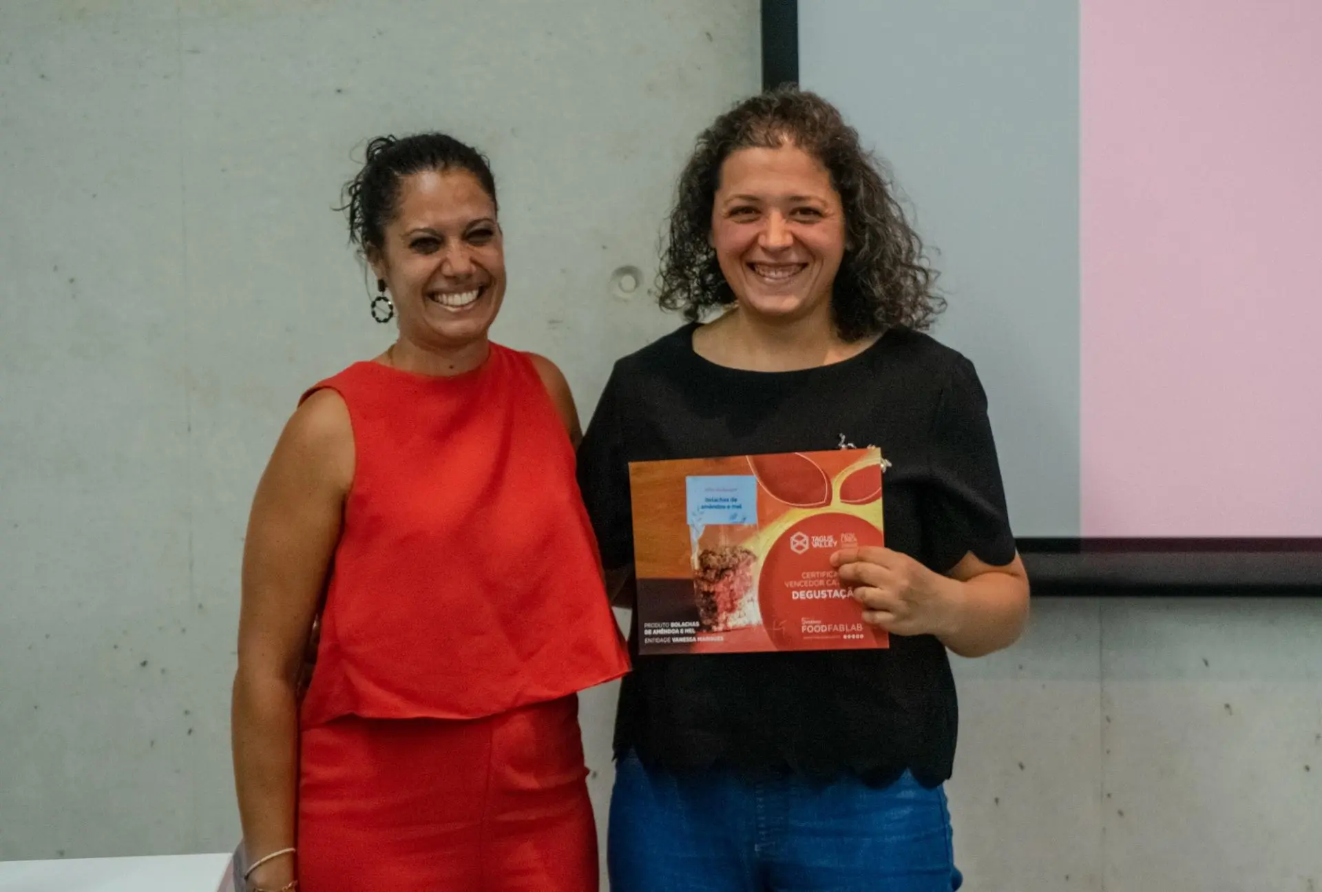 Duas mulheres sorridentes junto a quadro branco, uma com vestido vermelho, outra com blusa preta e calças azuis segurando caixa vermelha.