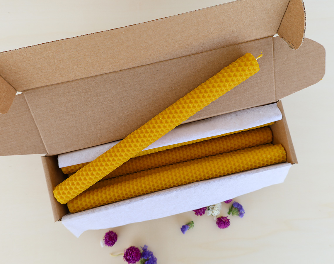 Velas amarelas de cera de abelha em caixa de papelão com forro branco e flores secas ao redor.