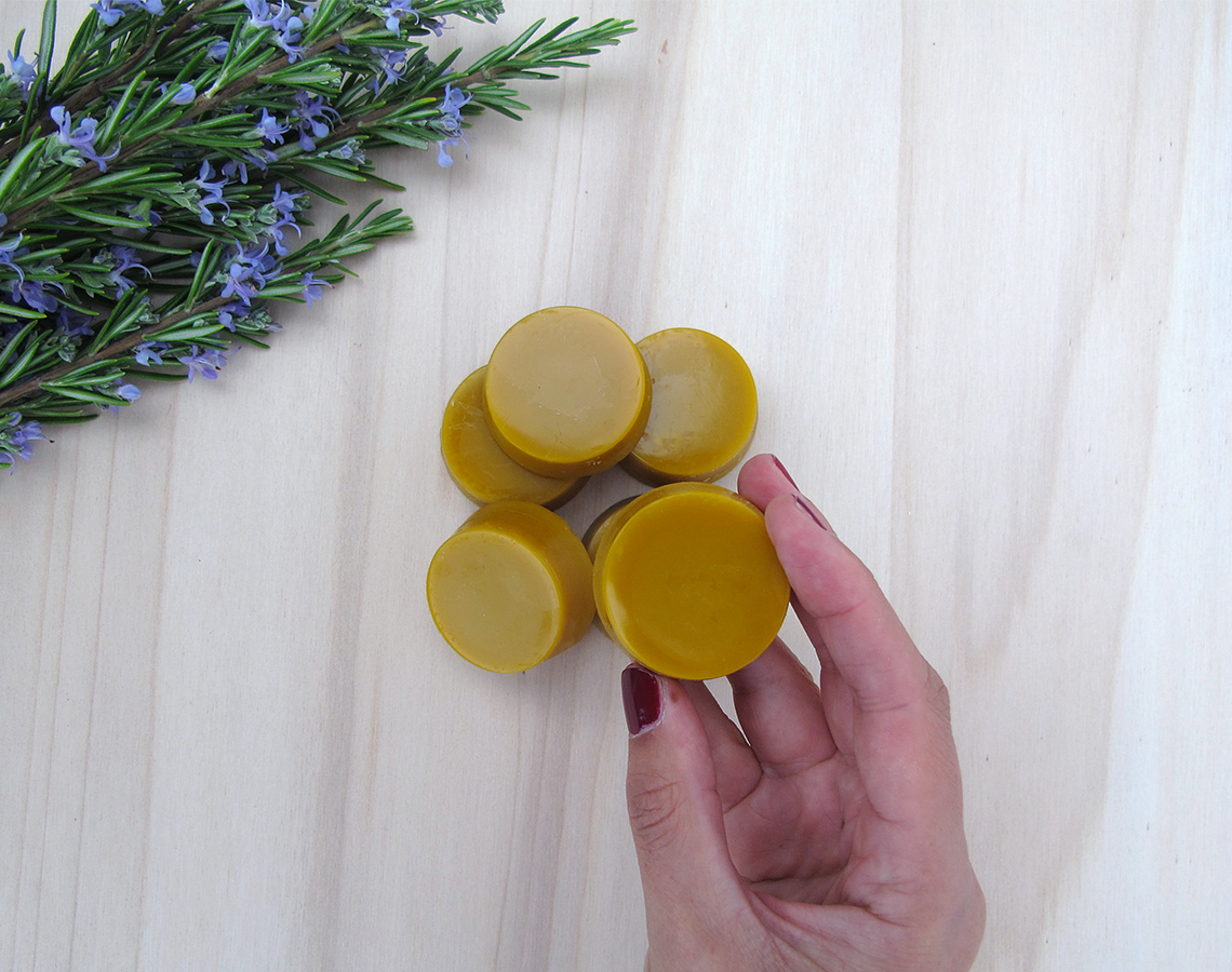 Bola de cera amarelo-dourada com uma mão segurando e erva com flores roxas sobre fundo de madeira clara