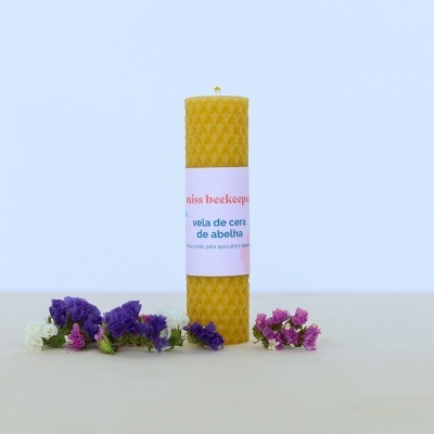 Vela cilíndrica de cera de abelha amarela com etiqueta branca e flores coloridas ao redor