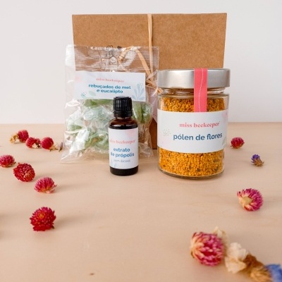 Conjunto de produtos de apicultura com pólen de flores, extrato de própolis e rebuçados de mel e eucalipto, com flores secas ao redor.