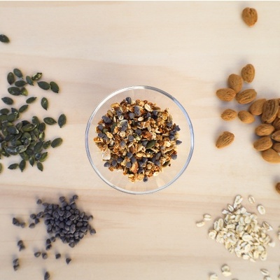 Tigela de vidro com granola cercada por amêndoas, pepitas de chocolate e sementes de abóbora sobre mesa de madeira