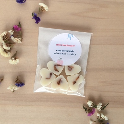 Pacote de corações de cera perfumada com flores secas e etiqueta sobre mesa clara, rodeado de flores secas coloridas.