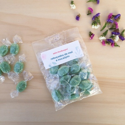 Pacote de rebuçados de mel e eucalipto com rebuçados verdes embalados individualmente em madeira clara, flores ao lado.