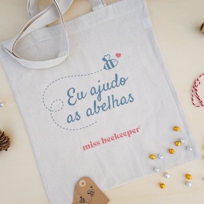 Saco de pano branco com texto e ilustração de abelha, em mesa clara com enfeites e fio